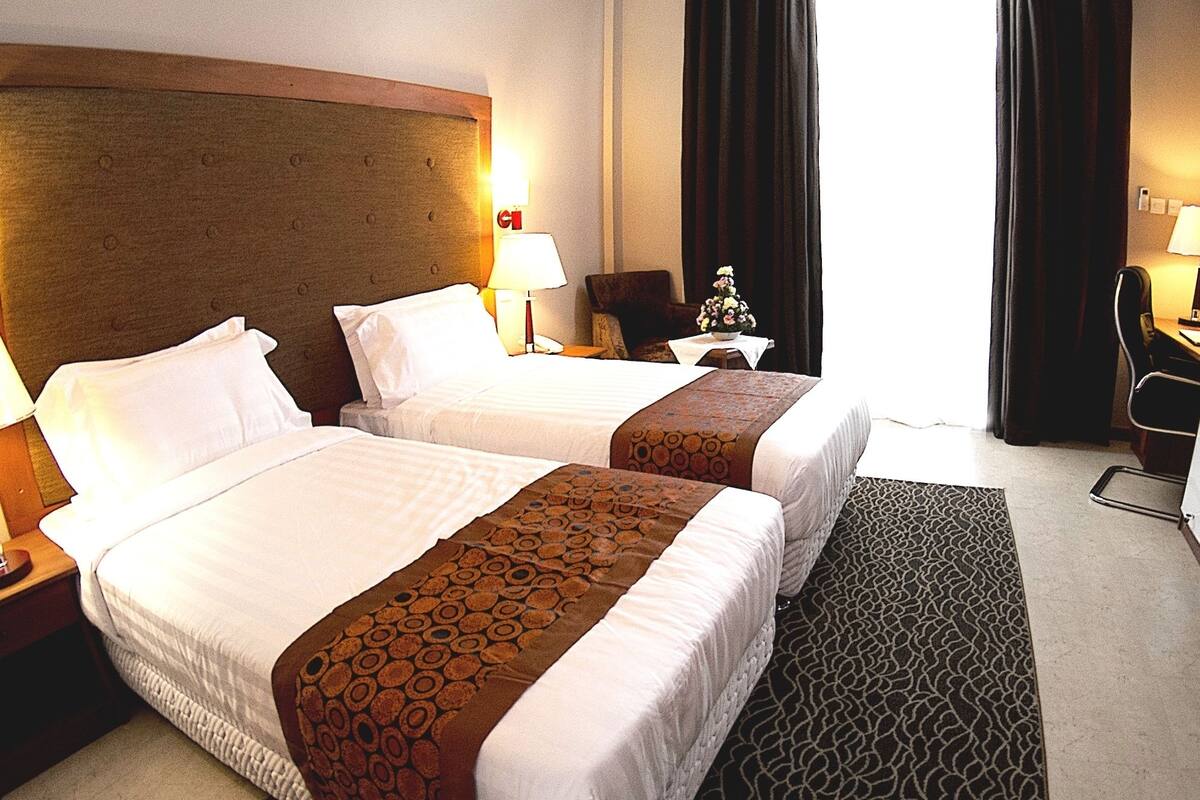 A hotel room with two beds, a desk, a chair, a TV, and a lamp.
