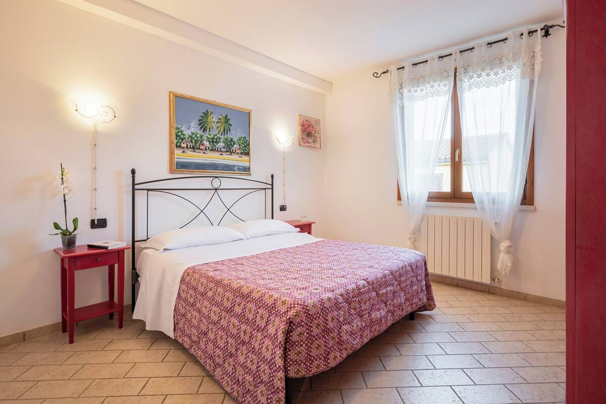A bedroom with a bed, a nightstand, a potted plant, a painting, and a window with sheer curtains.