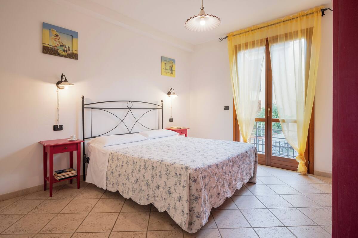 A bedroom with a bed, two bedside tables, a window with curtains, and a ceiling light.
