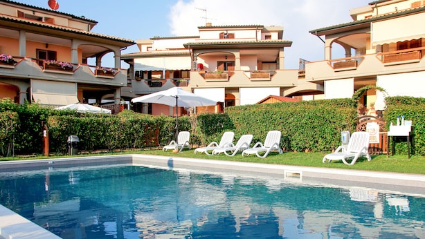 Piscina com espreguiçadeiras e guarda-sóis, cercada por sebes bem cuidadas e um edifício com varandas.