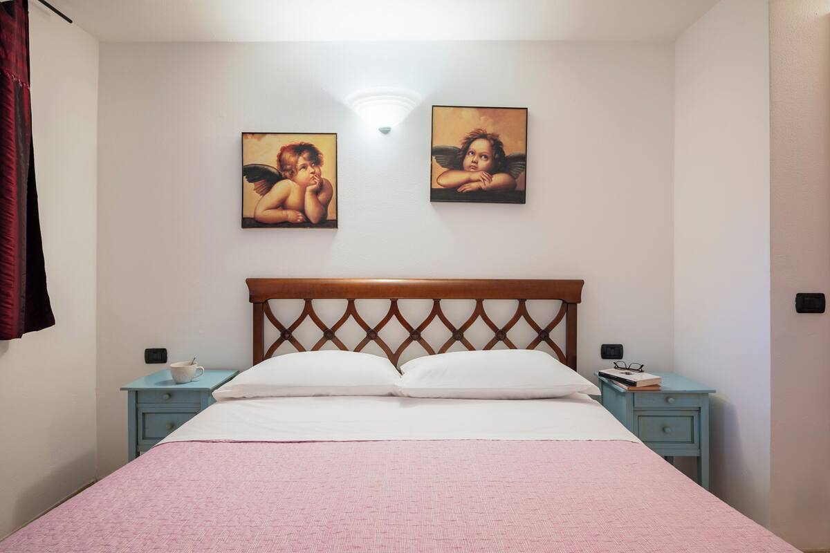 A bedroom with a wooden headboard, a bed with white linens, two nightstands with a cup and book on one, and two wall-mounted paintings.
