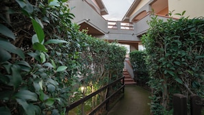 Uma passagem estreita ladeada por vegetação exuberante, conduzindo a um edifício com arquitetura moderna.