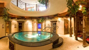 Een wellnessruimte met een centrale jacuzzi, stenen muren, houten balken en een balkon met een hekwerk.