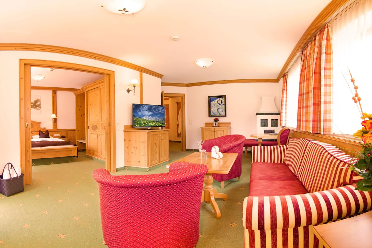 Une chambre d’hôtel avec un canapé, un fauteuil et une télévision. On y trouve également une armoire en bois et une table sur laquelle est posé un vase de fleurs.