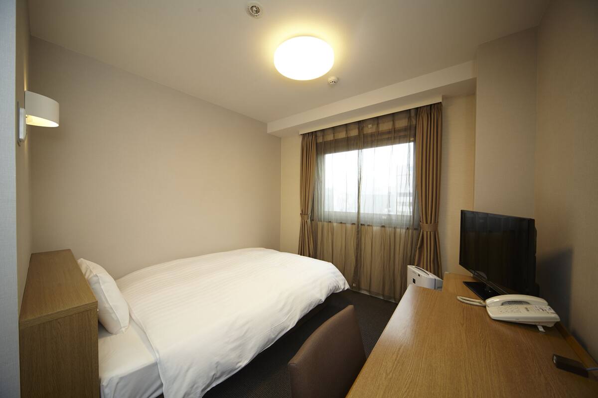 A hotel room with a single bed, a desk with a telephone and a television, and a window with curtains.