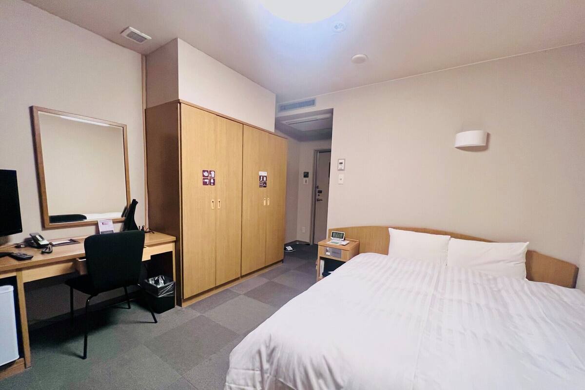 A hotel room with a bed, a desk with a computer, a chair, a wardrobe, and a nightstand with a lamp.