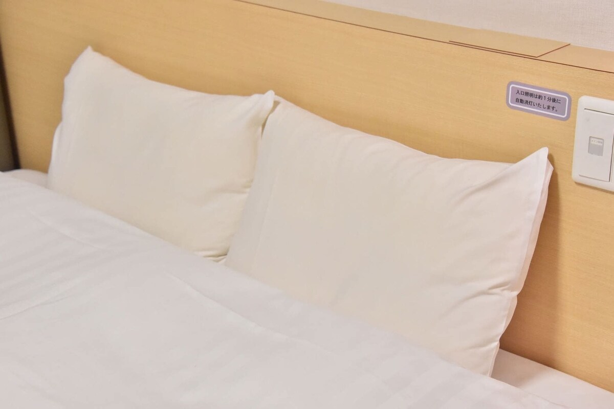 A bed with white bedding and pillows, a wooden headboard, and a wall-mounted light switch.