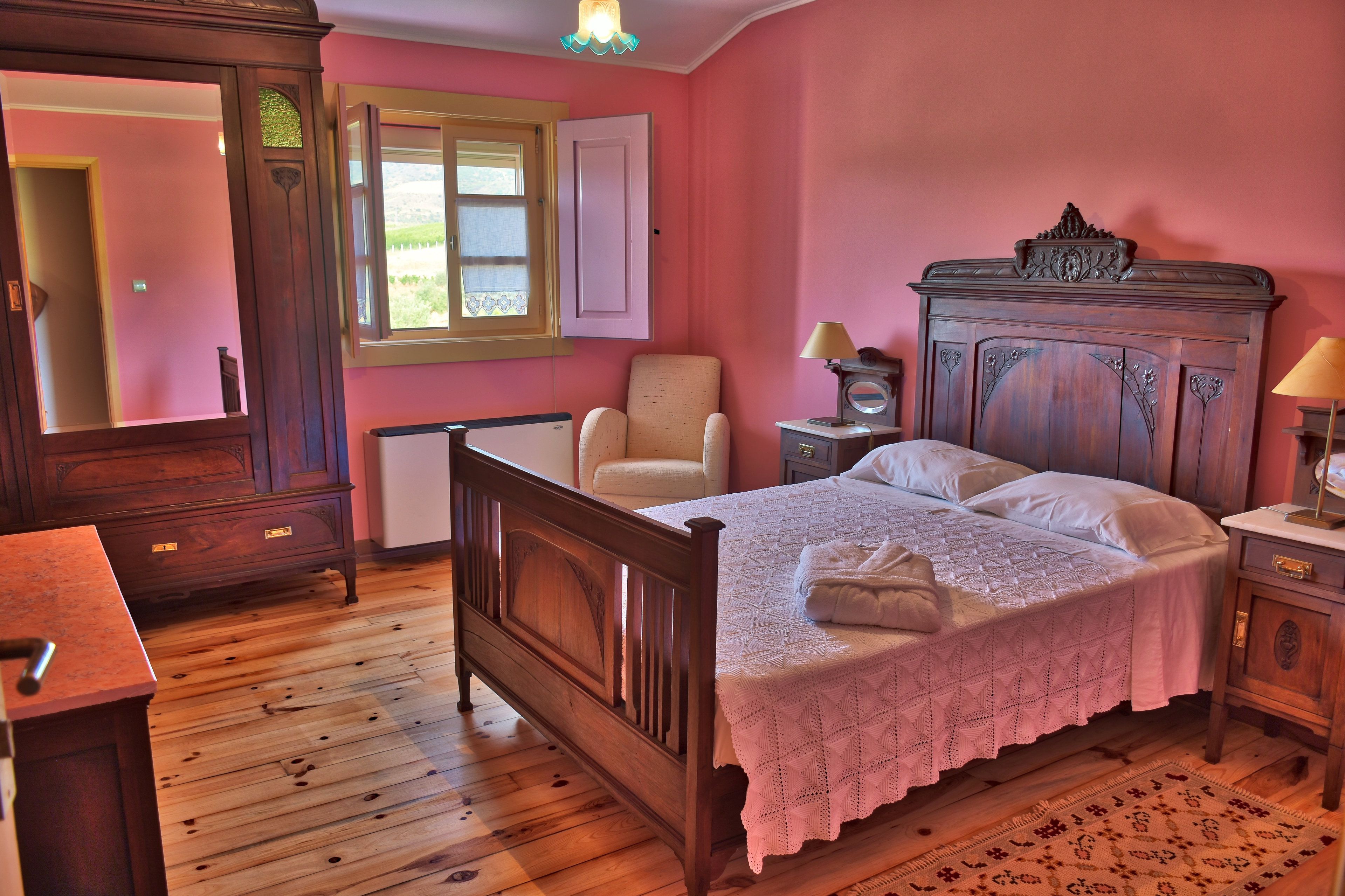 A bedroom with a wooden bed, a chair, a dresser, a nightstand, and a window with a view of greenery.