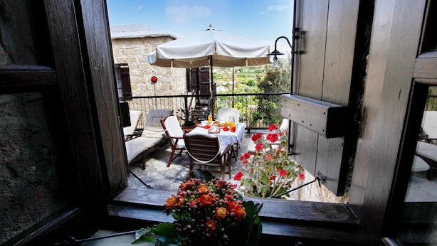 An open balcony with a table and chairs, a white umbrella, and a stone building in the background. - Leonidas Village Houses (Goudi)
