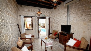 A cozy living room with stone walls, wooden furniture, a television, and a view of the outside. - Leonidas Village Houses (Goudi)