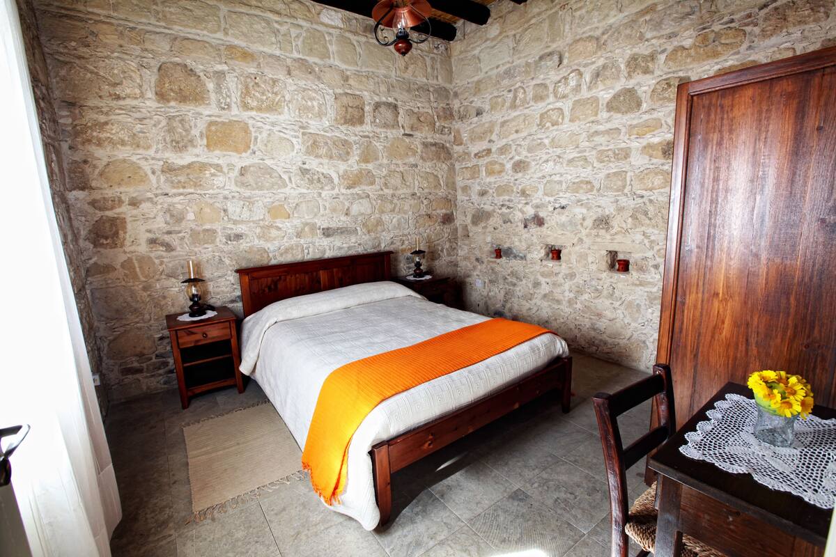 A stone-walled bedroom with a wooden bed, a nightstand with a lamp, a wooden chair, and a table with a vase of flowers.