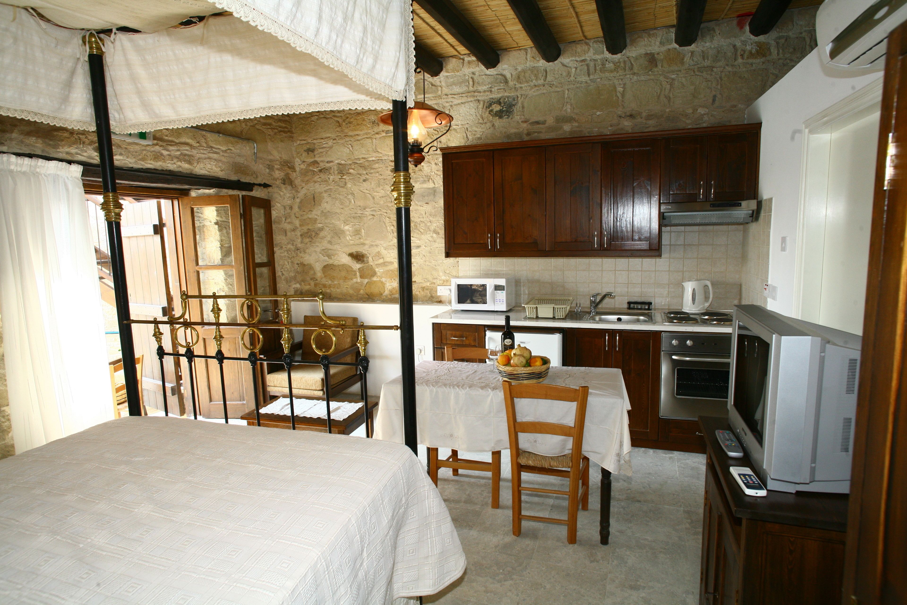 A rustic kitchen with wooden cabinets, a dining table with chairs, a bed with a white bedspread, and a small table with a bowl of fruit.