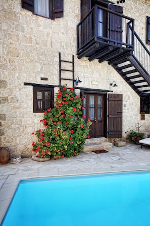 A stone building with a wooden staircase, a pool, and a bush with red flowers.