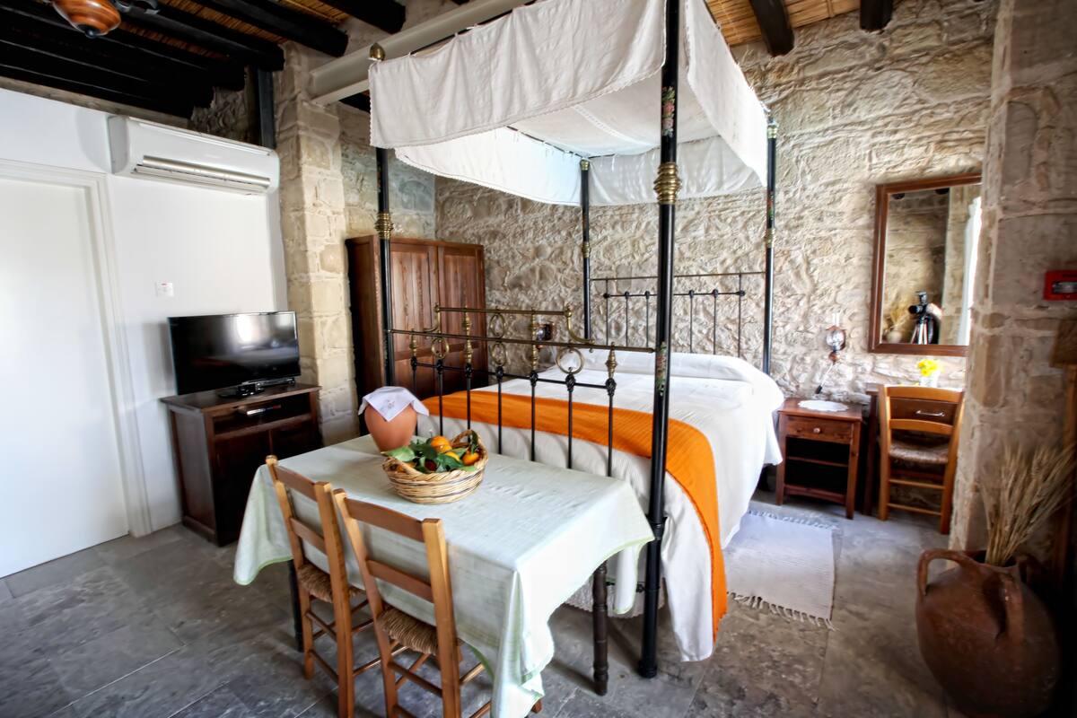 A bedroom with a stone wall, a bed with an orange bedspread, a wooden nightstand, a television, and a wicker basket on the table.