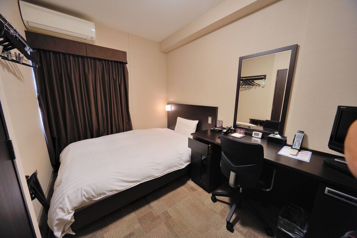 A hotel room with a bed, a desk, a chair, a desk lamp, a telephone, a mirror, and a television.