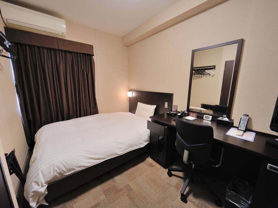 A hotel room with a bed, a desk, a chair, a desk lamp, a telephone, a mirror, and a television.