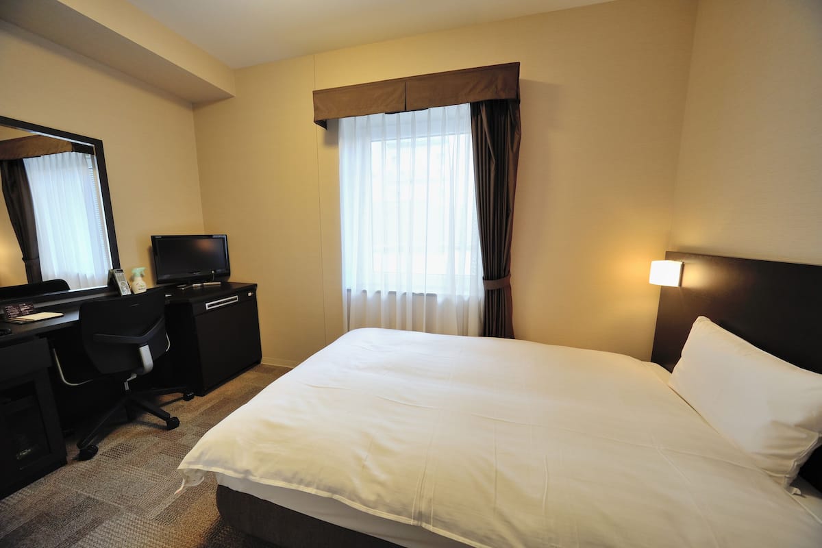 A hotel room with a bed, a desk with a computer, a chair, and a window with curtains.