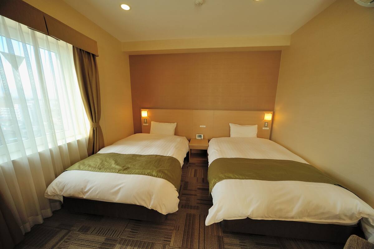 A hotel room with two beds, a large window with curtains, a headboard with a light, and a carpeted floor.