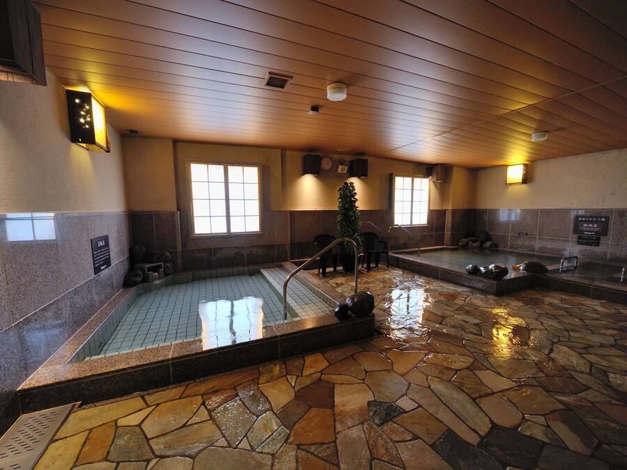 A stone-floored indoor pool area with two separate pools, wooden ceiling, and wall-mounted lighting fixtures.