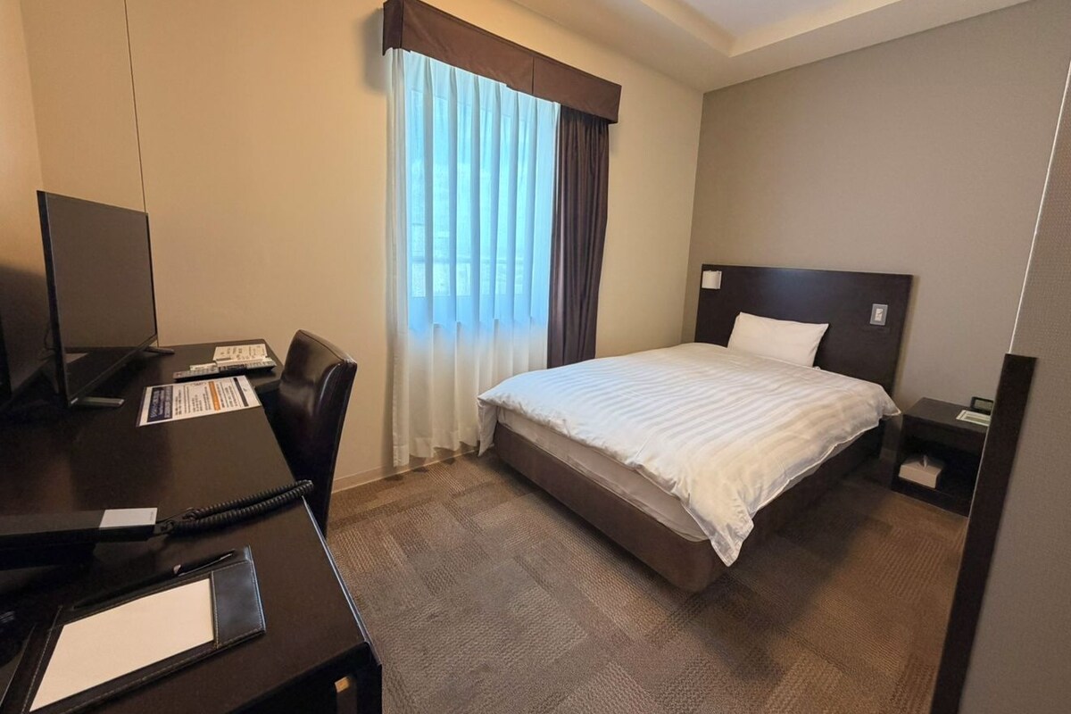 A hotel room with a bed, a desk with a telephone, a chair, and a nightstand with a lamp.