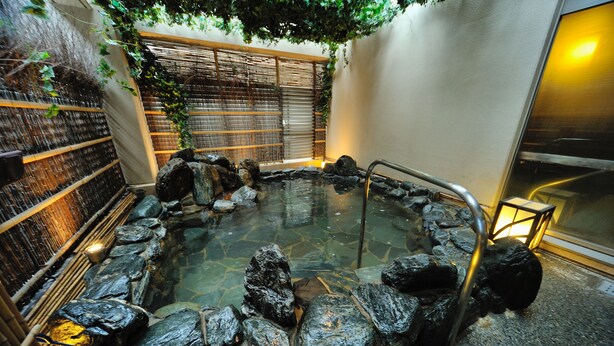 Un área de aguas termales con una piscina de piedra rodeada de rocas, una valla de madera y una pared de bambú.