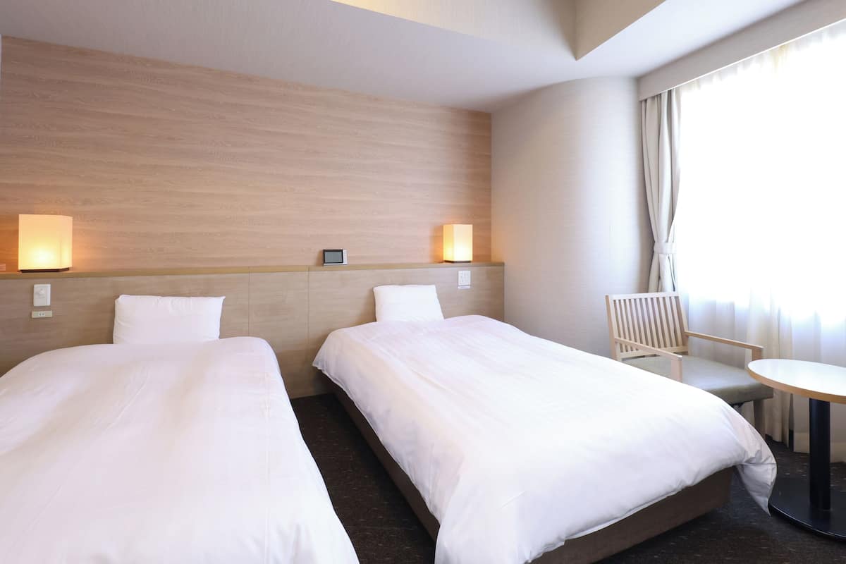 Ein Hotelzimmer mit zwei Betten, einem Sessel, einem kleinen Tisch und einem Fenster mit Vorhängen.