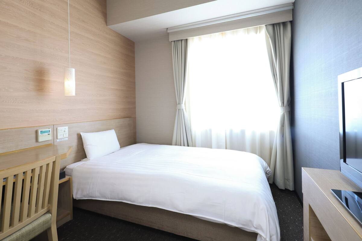Ein Hotelzimmer mit Bett, einem Holztisch, einem Fernseher und einem Fenster mit Vorhängen.