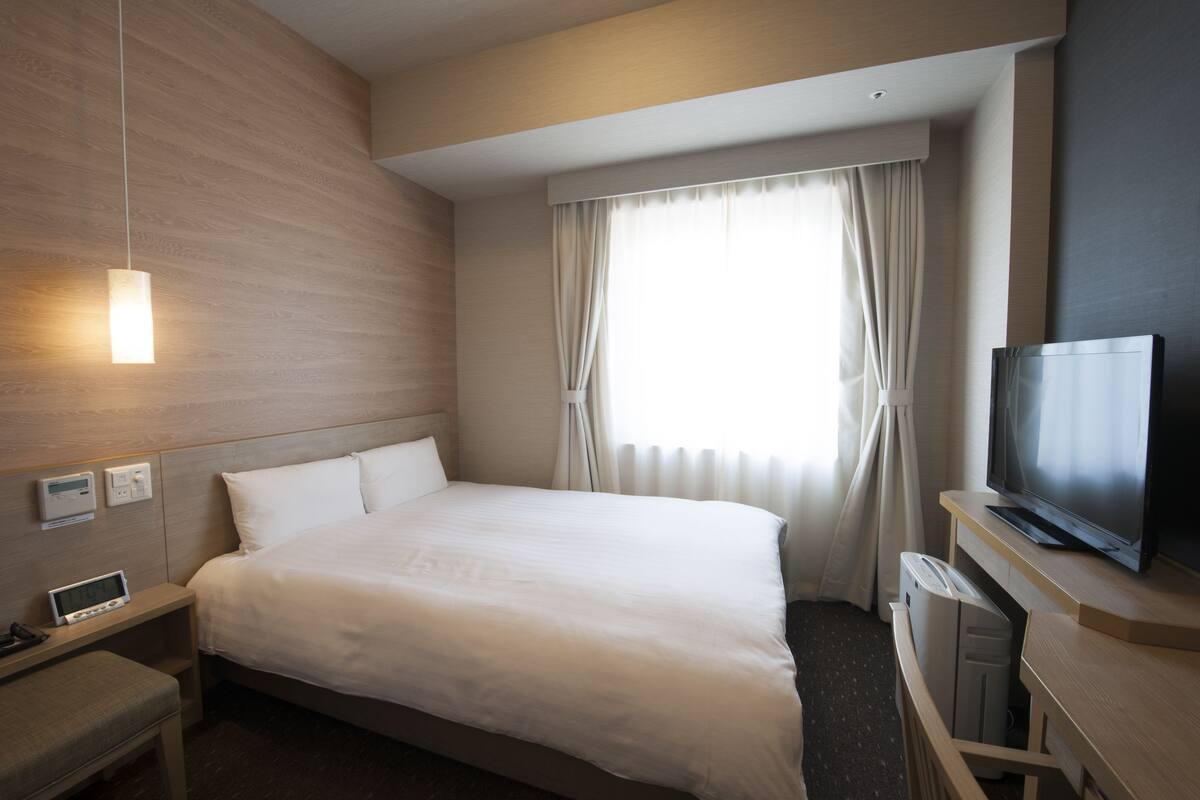 Ein Hotelzimmer mit einem großen Bett, einem Flachbildfernseher, einem Nachttisch mit Lampe und einem Fenster mit Vorhängen.