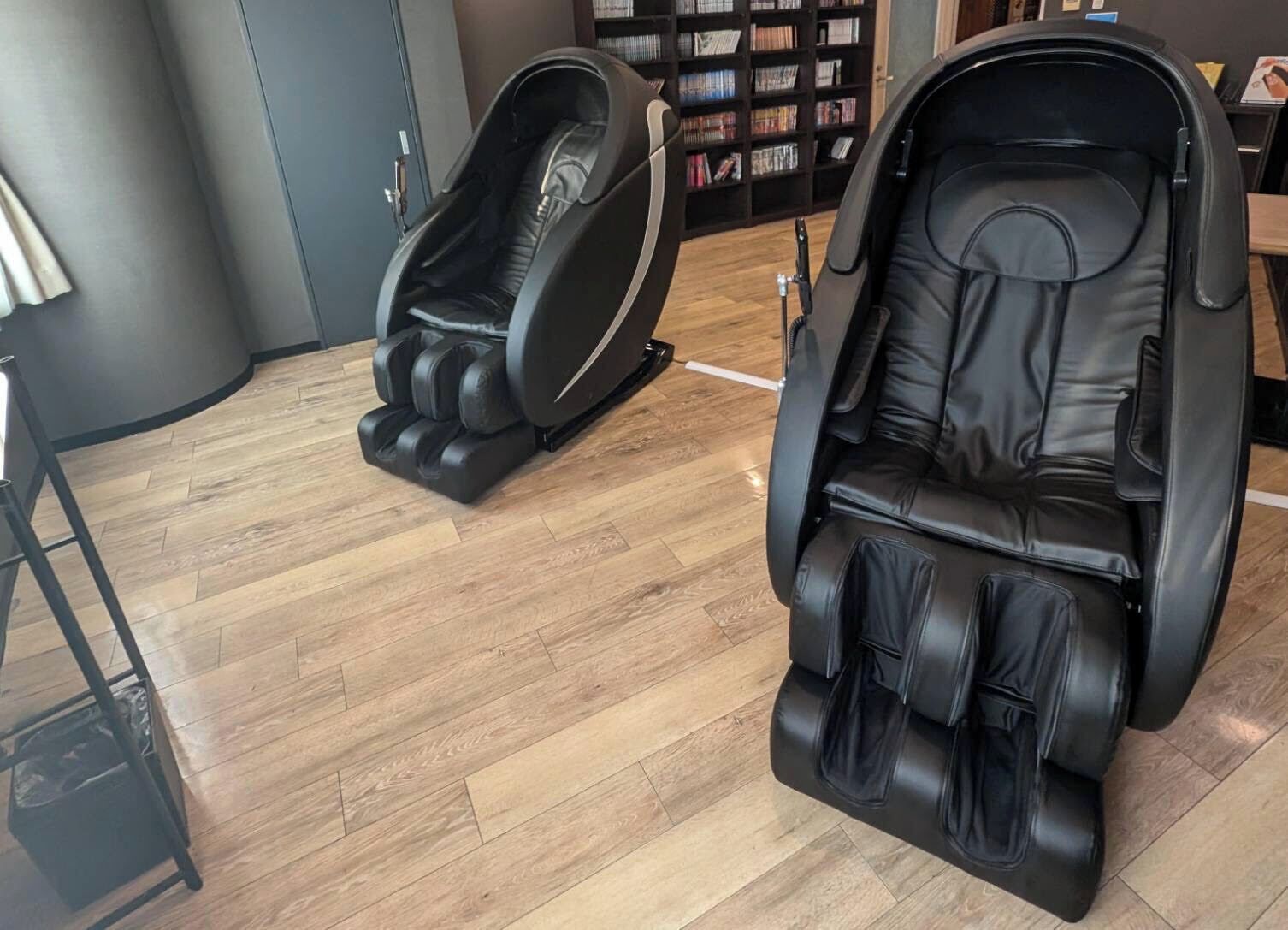 Two massage chairs in a room with wooden flooring and a bookshelf in the background.