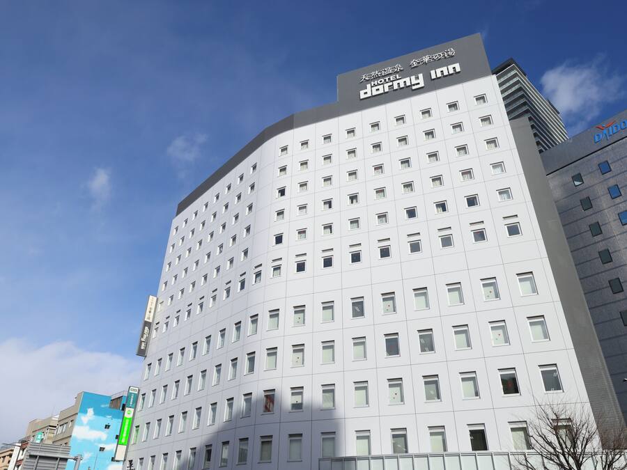 A modern hotel building with a prominent 'Hotel Dormy Inn' sign, featuring a grid of windows and a flat facade.