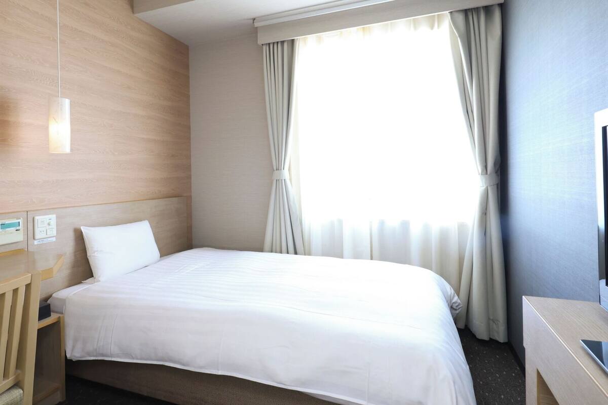 Ein Hotelzimmer mit einem Bett, Bedienelementen am Nachttisch, einem Fernseher und einem Fenster mit Vorhängen.