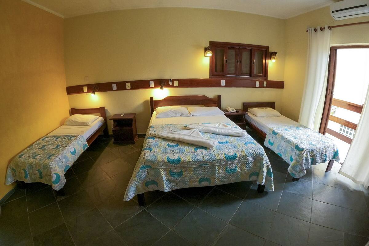 Ein Hotelzimmer mit drei Einzelbetten, jeweils mit einem gemusterten Überwurf, einem Holz-Kopfende und Nachttischlampen. Über den Betten befindet sich ein Holzschrank und an der Wand eine Klimaanlage.