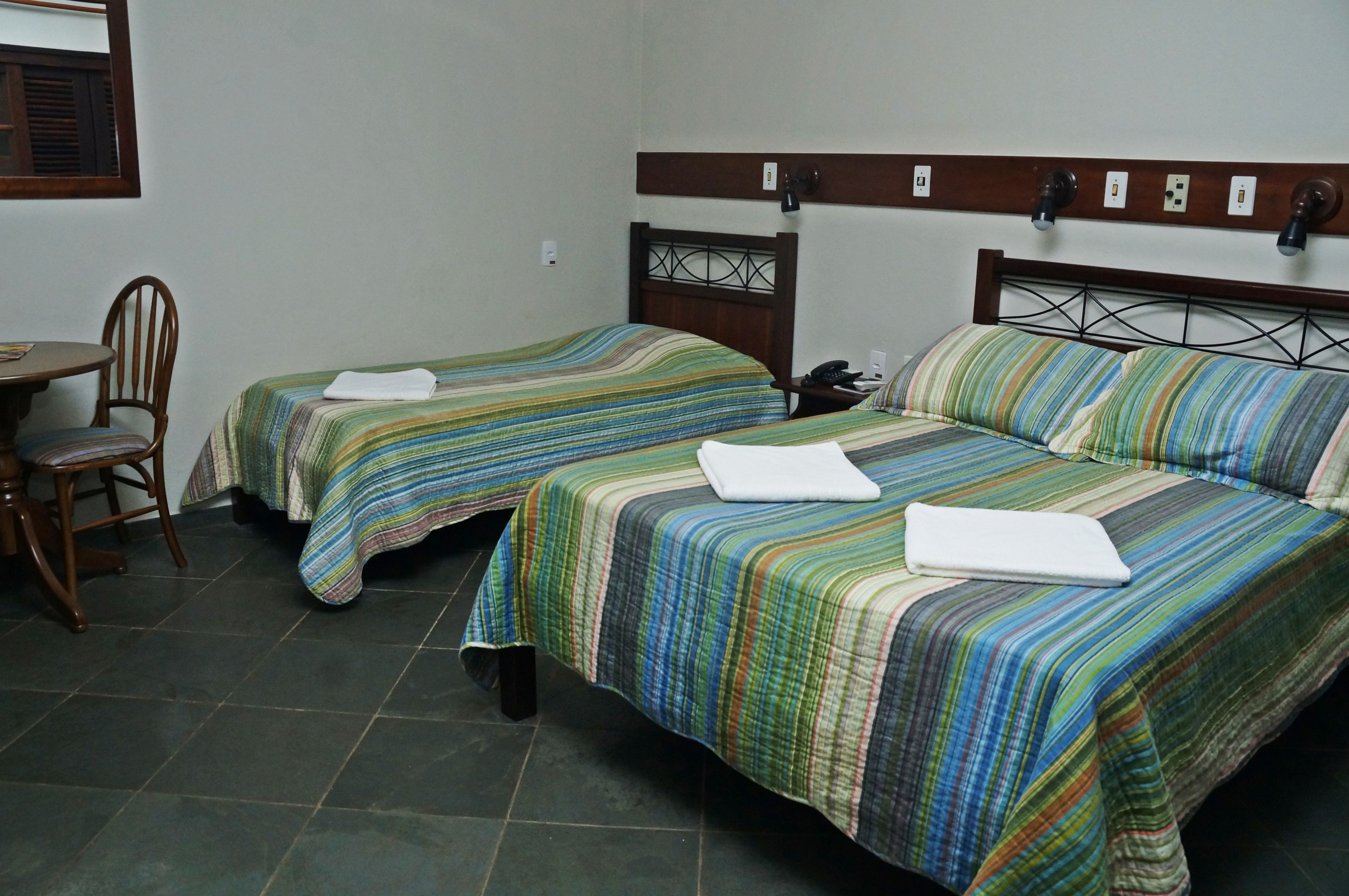 Une chambre d’hôtel avec deux lits simples, une tête de lit en bois, une petite table avec une chaise et une fenêtre avec des stores.