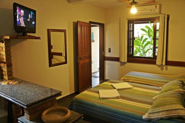 Quarto de hotel com cama, televisão, escrivaninha e janela vista para vegetação.