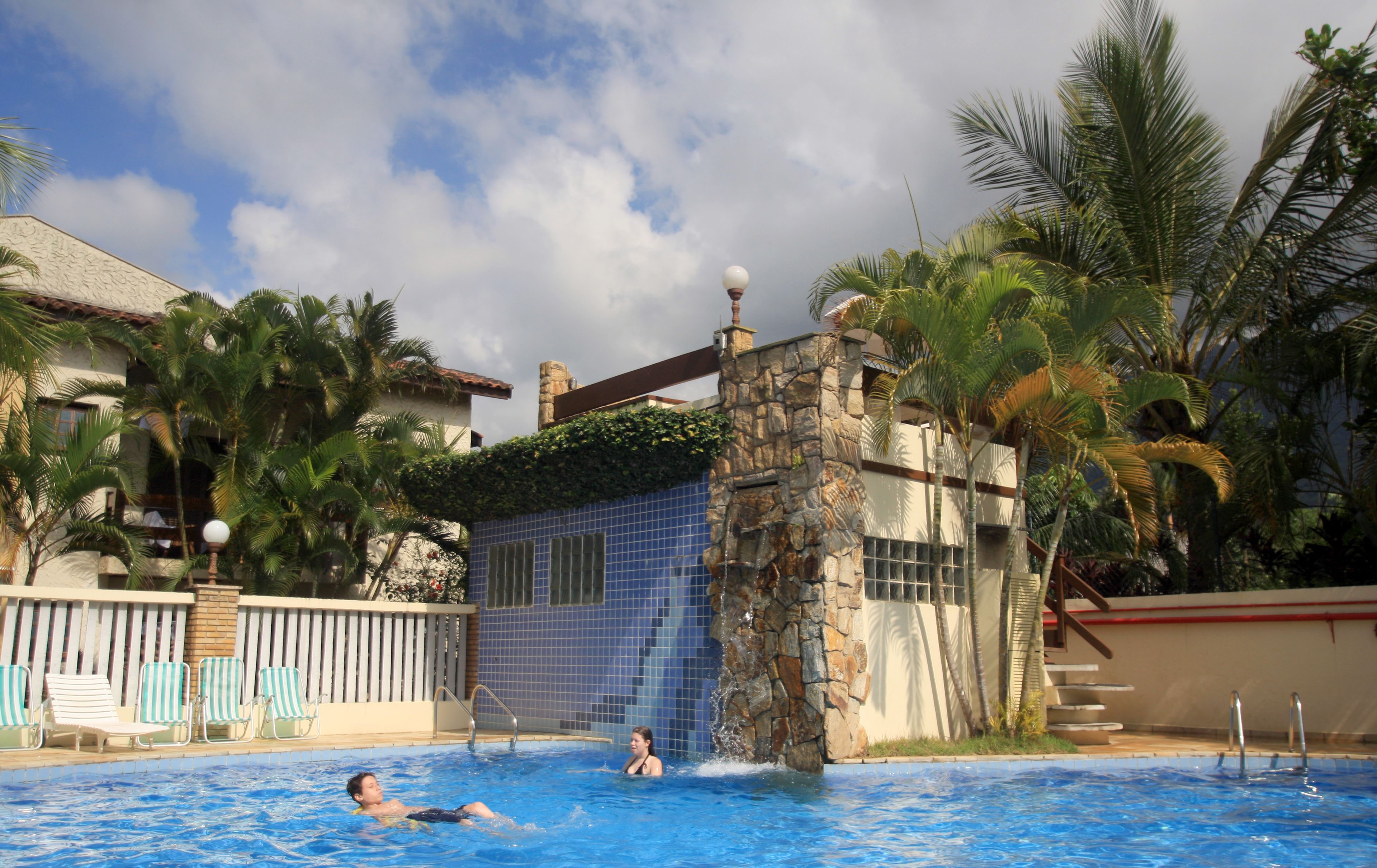Piscina com uma cascata de pedra, rodeada por palmeiras e um muro de azulejos.