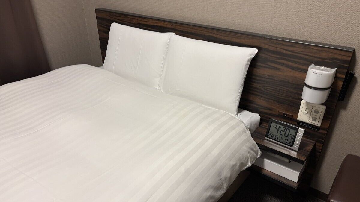 Ein Hotelzimmer mit Bett, weißen Leinentextilien, einem hölzernen Kopfteil, einem Nachttisch mit digitaler Uhr und einer Kaffeemaschine.
