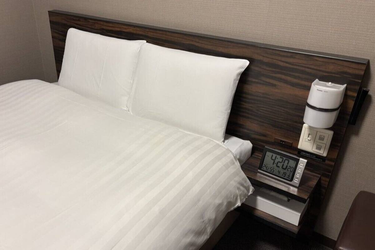 Habitación de hotel con cama, ropa de cama blanca, cabecero de madera, mesita de noche con reloj digital y cafetera.