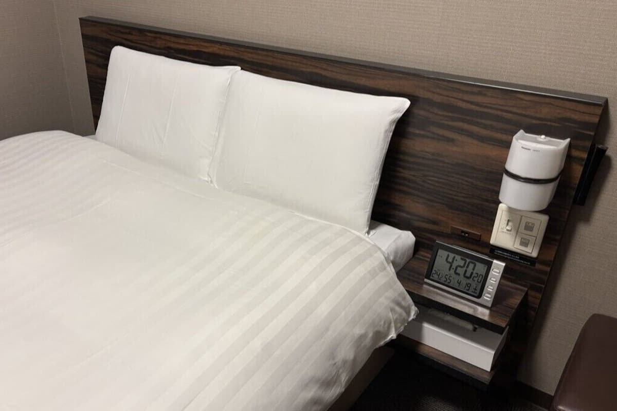 Ein Hotelzimmer mit Bett, weißen Leinentextilien, einem hölzernen Kopfteil, einem Nachttisch mit digitaler Uhr und einer Kaffeemaschine.
