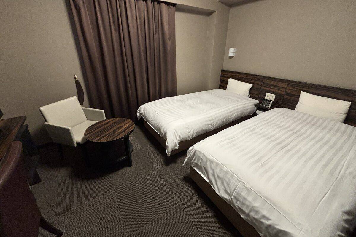 Habitación de hotel con dos camas individuales, un escritorio con silla, una mesita redonda y un gran ventanal con cortinas.