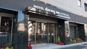 En modern hotellentrén med stora glasdörrar, en skylt med hotellets namn och en liten trädgårdsyta med röda blommor.