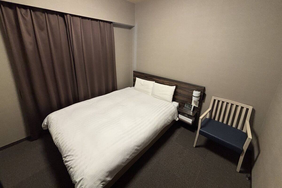 Ein Hotelzimmer mit einem Einzelbett, einem Holfnachtkästchen, einem Stuhl und einem Fenster mit Vorhängen.