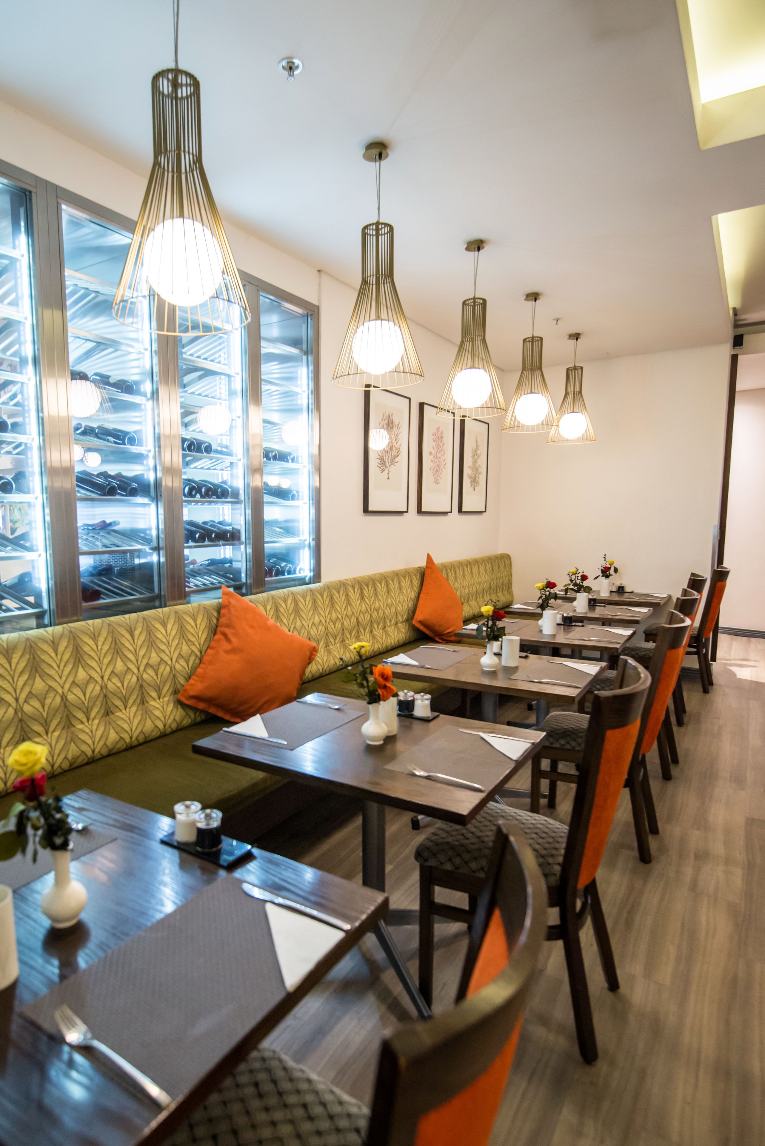 Un restaurante con una larga mesa de comedor, numerosas sillas y un sofá con cojines naranja. Hay lámparas colgantes sobre la mesa, y las paredes están decoradas con cuadros enmarcados.