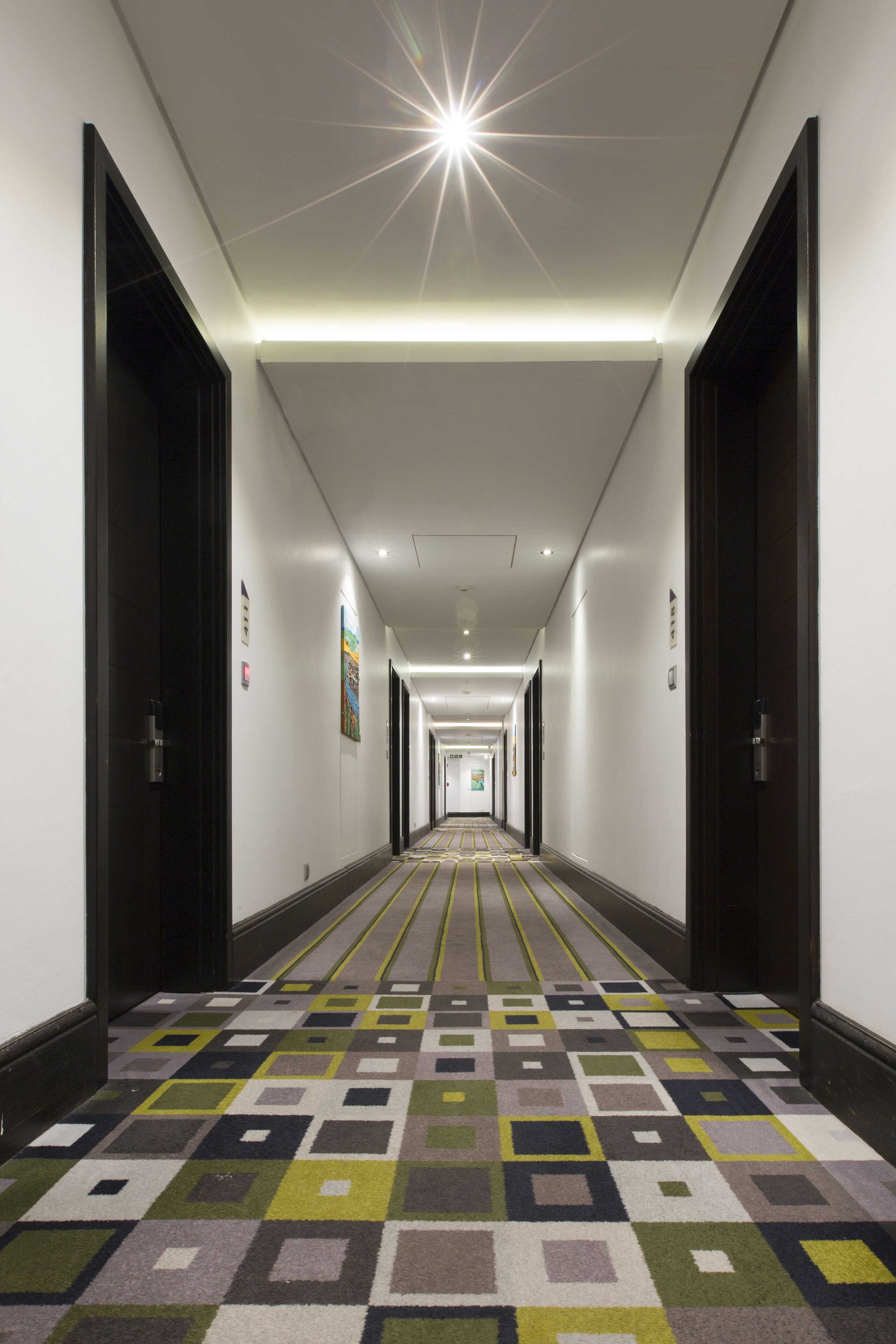 Un couloir d’hôtel avec un tapis à motifs, des portes sombres et un luminaire en forme d’étoile au plafond.