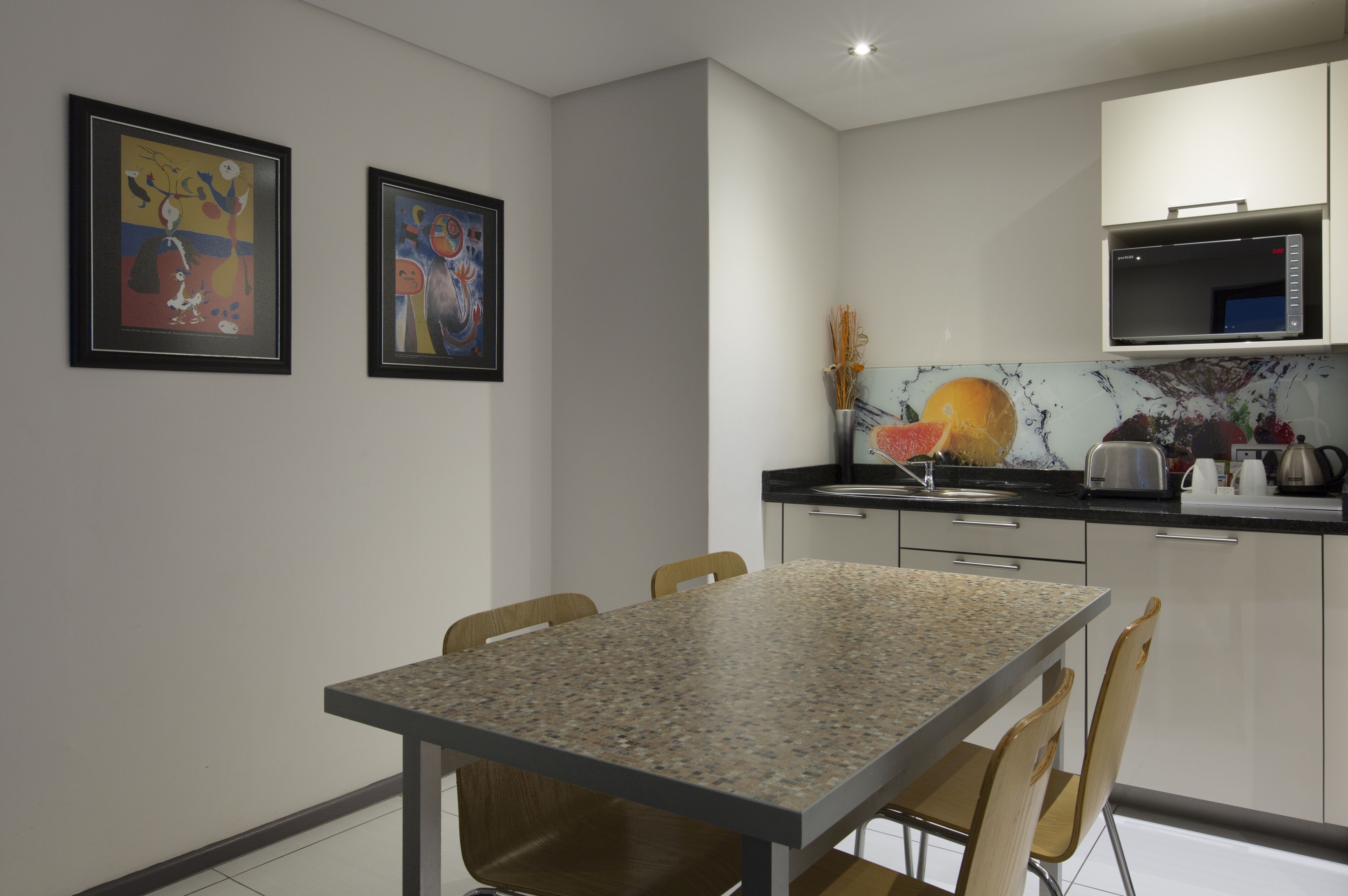 A modern kitchen with a granite countertop, white cabinets, and a microwave. There are two framed abstract paintings on the wall.
