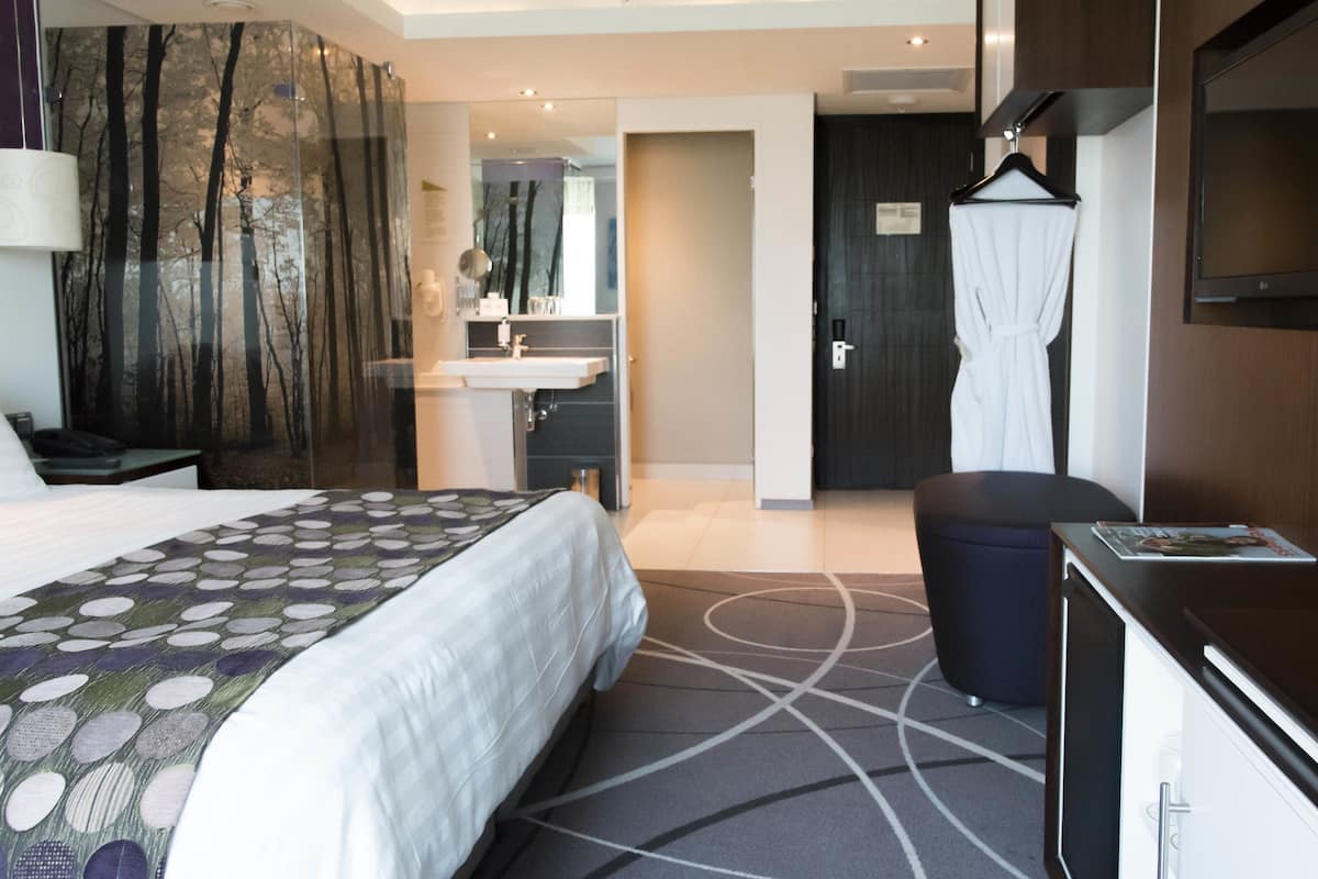 Une chambre d’hôtel avec un grand lit, un bureau, un miroir, une armoire et une salle de bain visible à travers une porte ouverte.