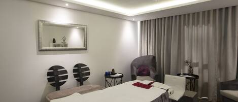 A modern hotel room with a massage table, a chair, a small table with toiletries, and a large mirror on the wall.