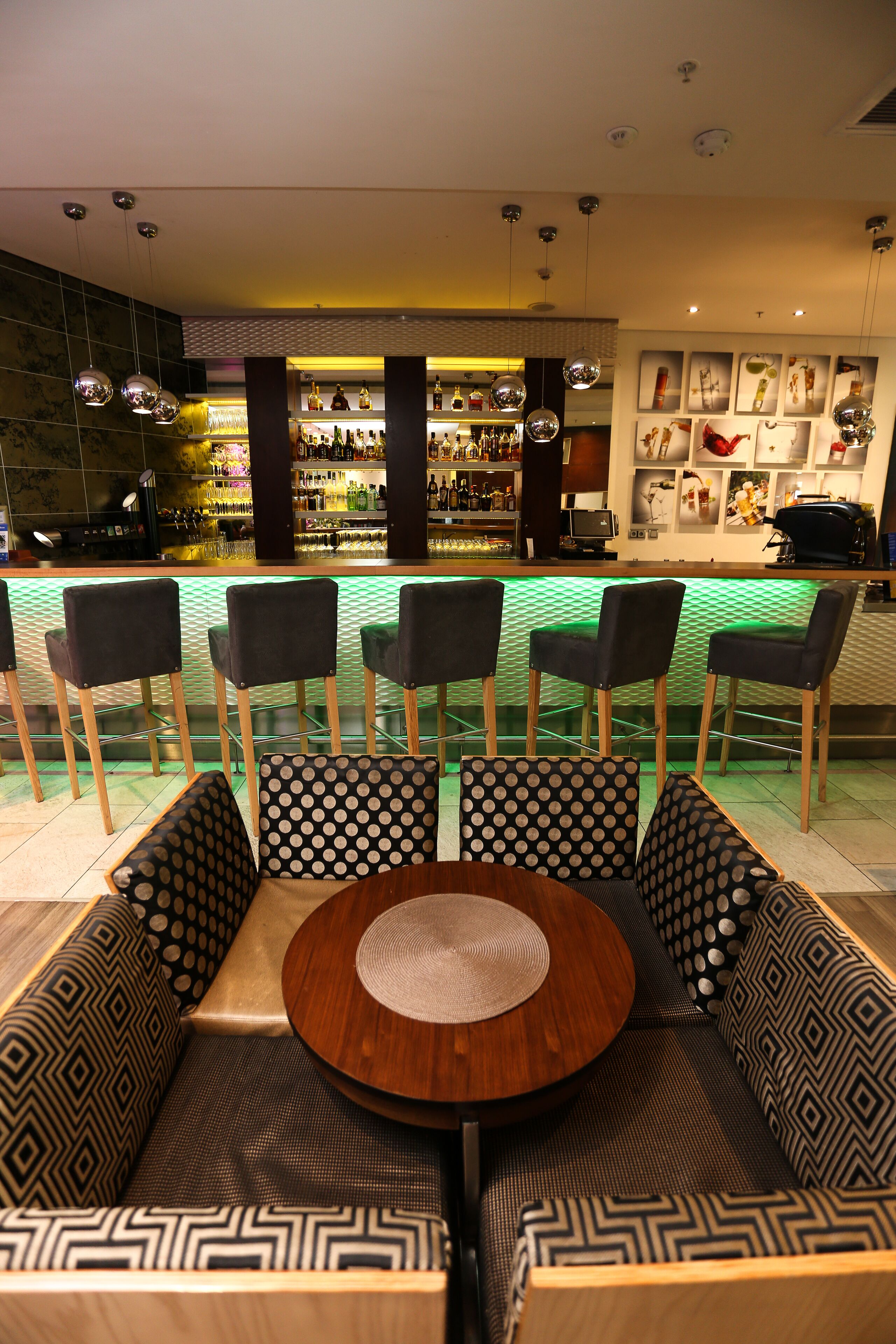 A bar area with a seating arrangement of five chairs and a sofa, a round wooden table, and a patterned wall with framed pictures.