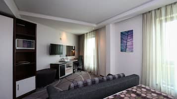 A modern hotel room with a bed, a sofa, a desk with a computer, a television, and a large window with curtains.