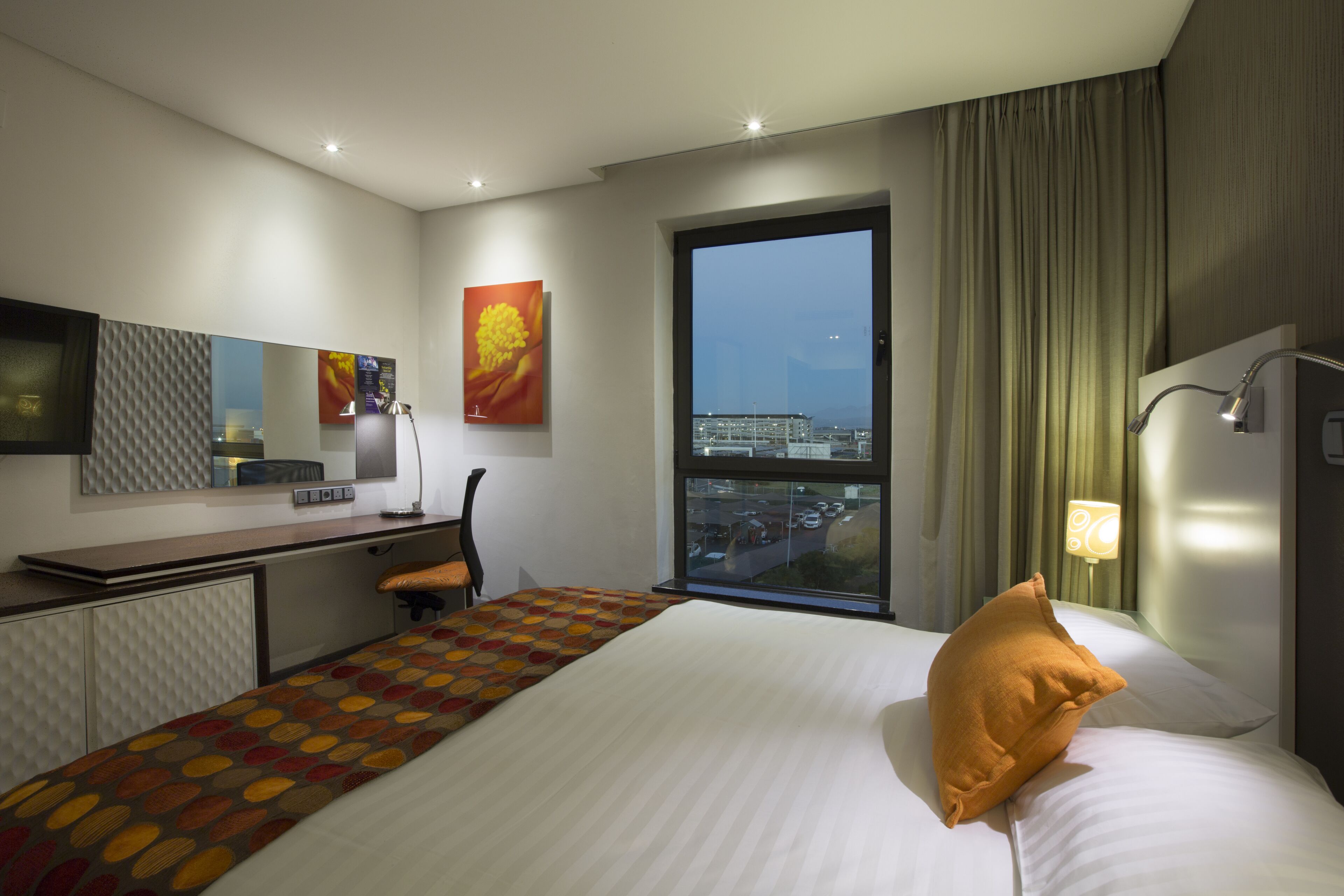 A hotel room with a large bed, a desk with a chair, a window with a city view, and a wall-mounted TV.