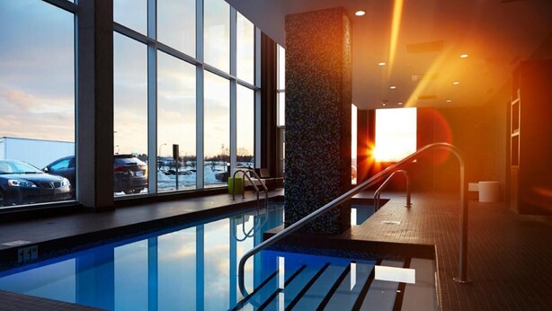 An indoor swimming pool with clear glass walls, a view of parked cars outside, and a sunset visible through the windows. - Alt Hotel Halifax Airport - In Terminal (Goffs)